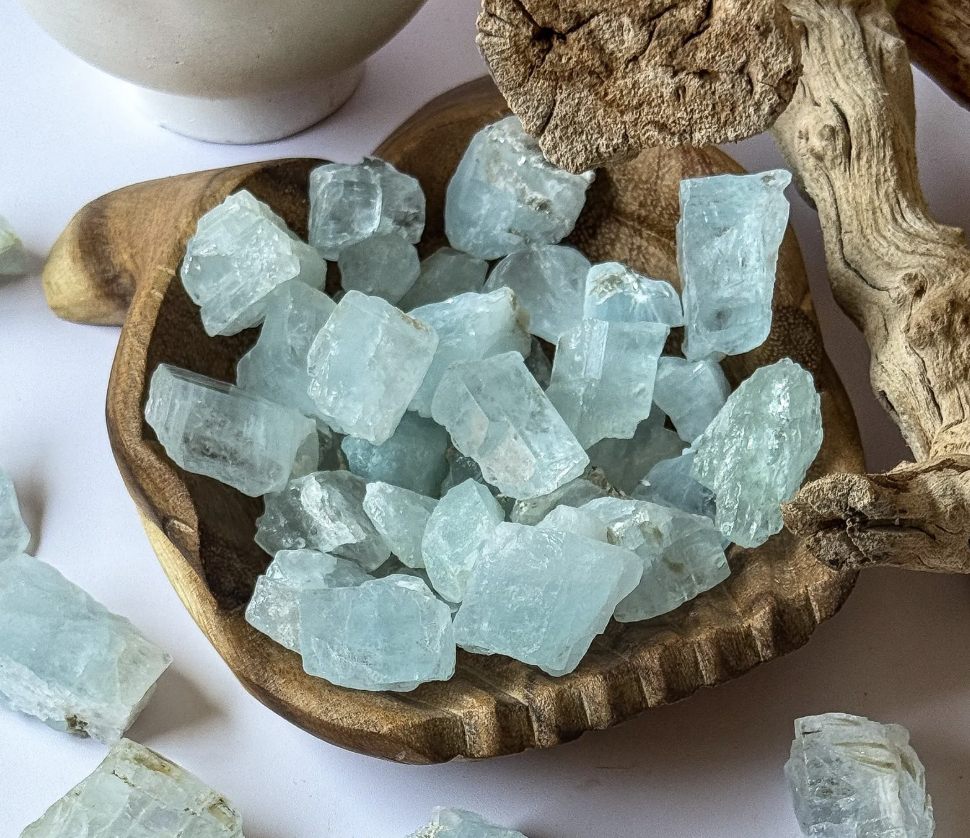 Raw pale blue Aquamarine stones displayed in a carved wooden bowl, used for balancing Aries energy, with driftwood and ceramic elements nearby.